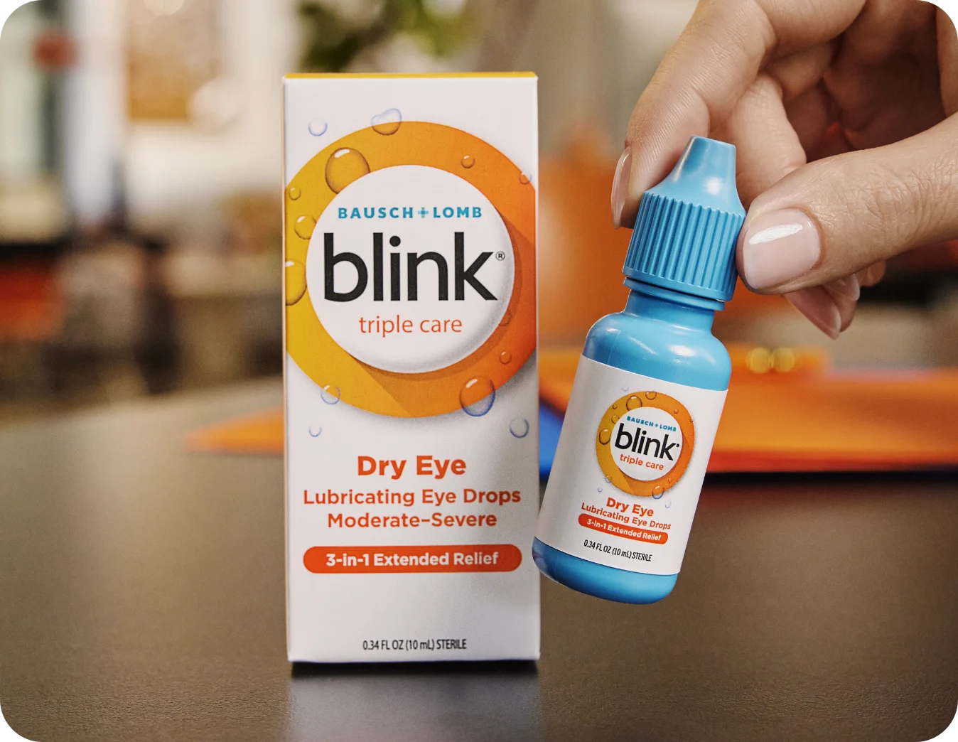 A person sitting at a kitchen table applying lubricating eye drops, with a calm neutral expression and soft natural window light