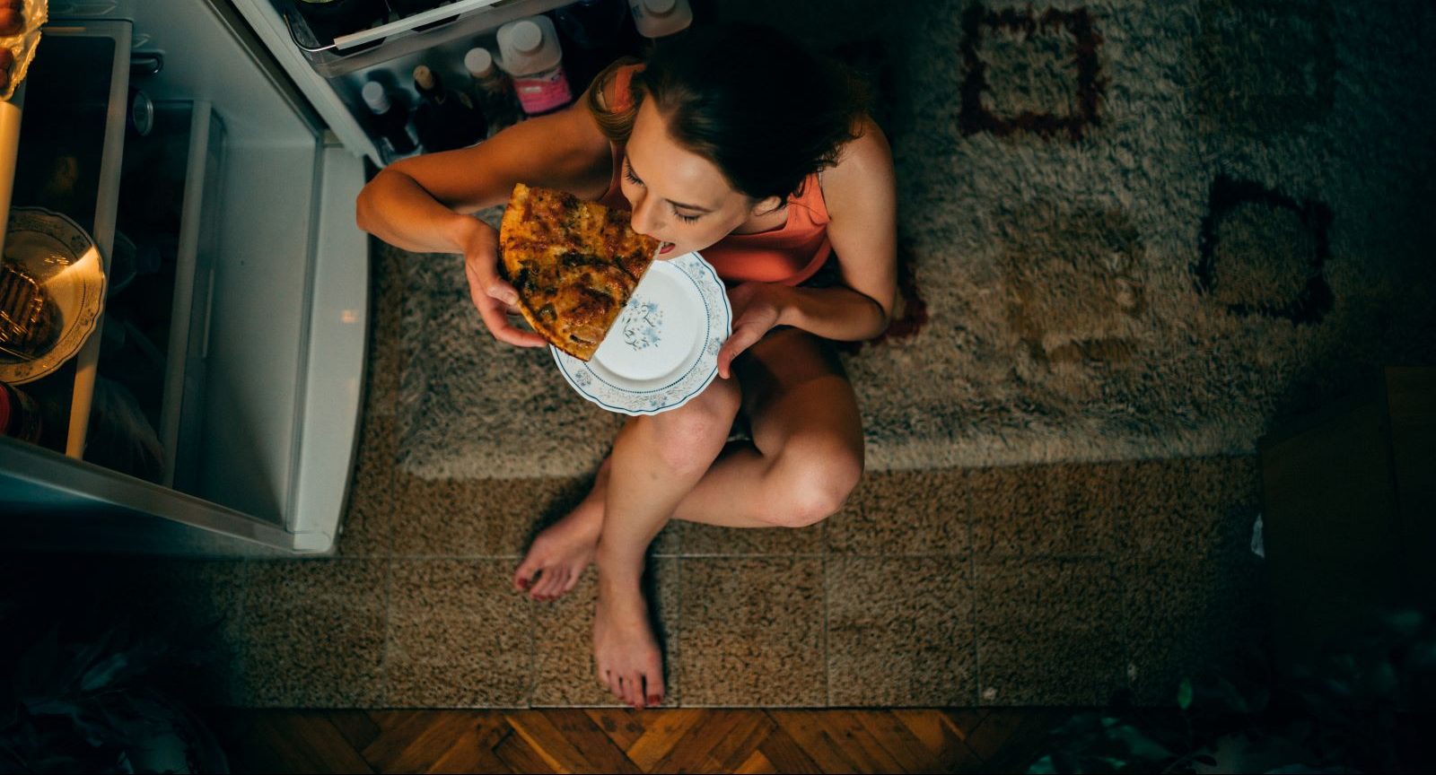 A person sitting at a kitchen table late at night with a large plate of food and a beverage, appearing tired and about to eat close to bedtime