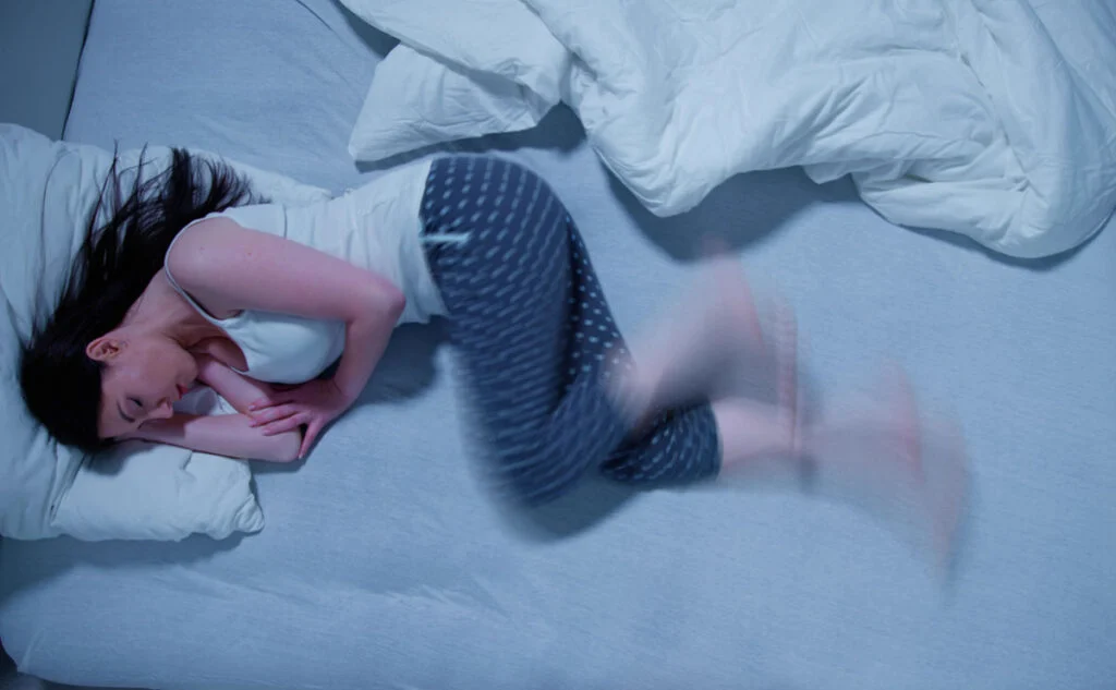 A tired adult sitting on the edge of a bed at night, rubbing their lower legs as if uncomfortable and unable to settle, with a dim bedside lamp in the background