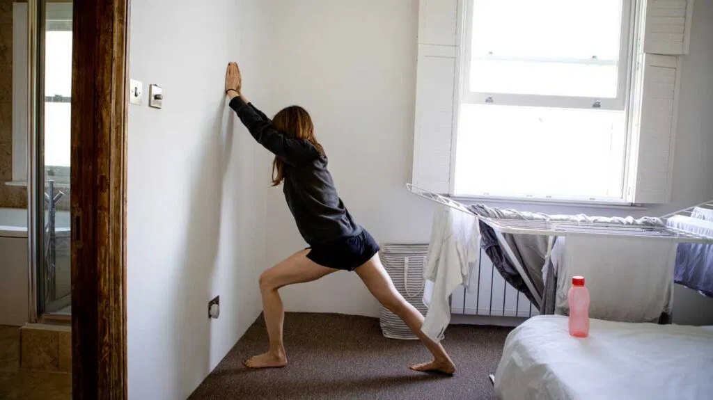 An adult doing a gentle calf stretch near the side of a bed in a softly lit bedroom, preparing for sleep