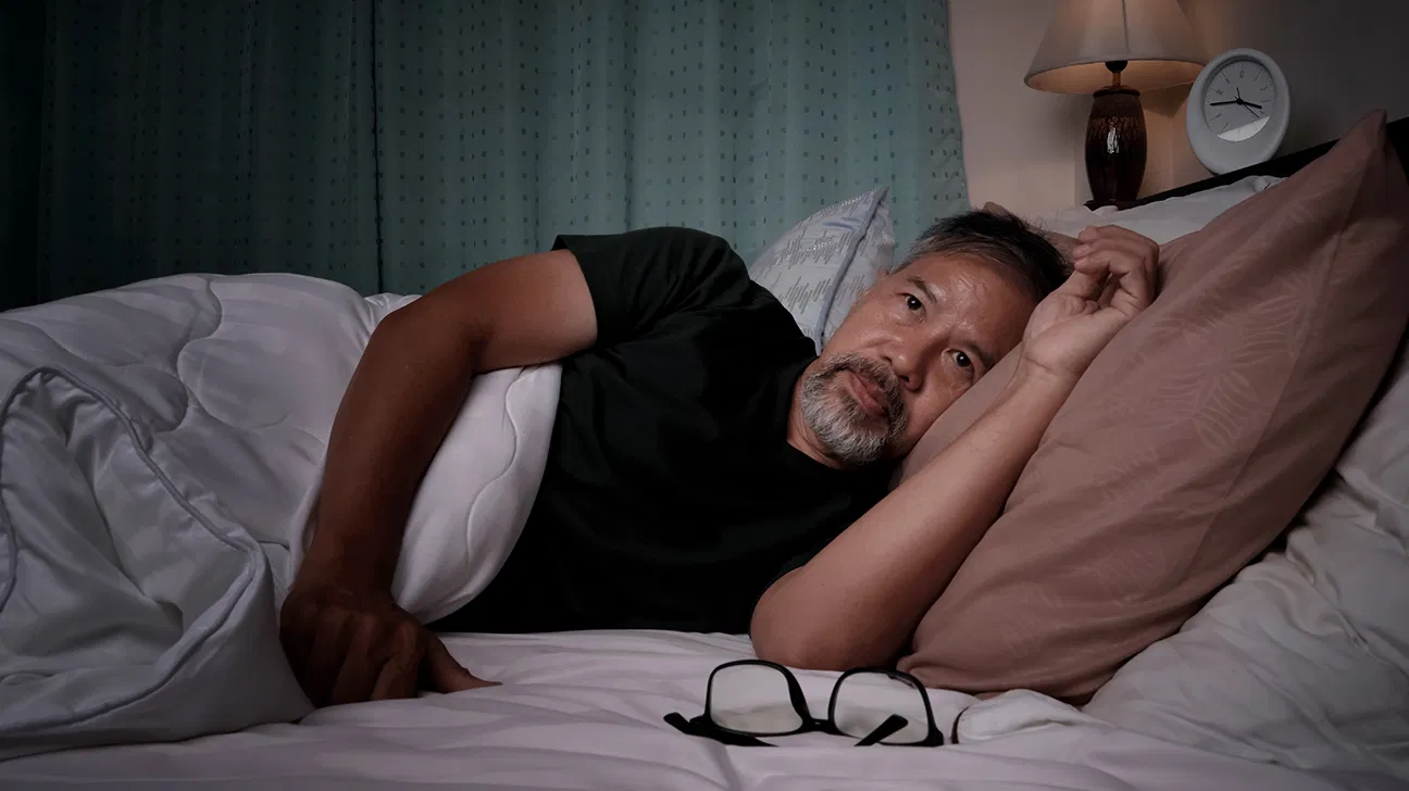 An adult lying awake in bed at night with a bedside lamp off, looking tired and thoughtful in a dark, quiet bedroom
