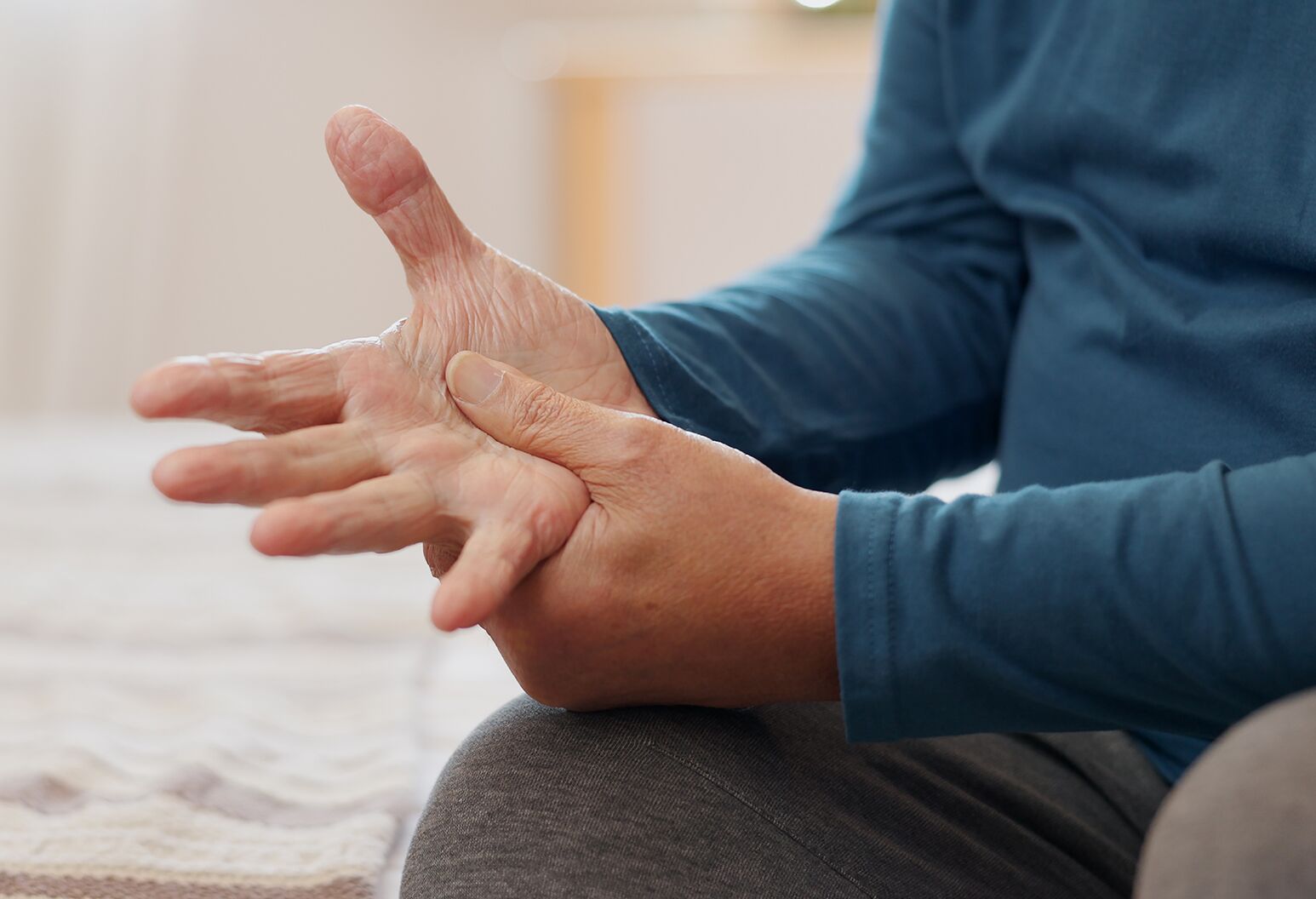 An adult sitting on the edge of a bed in morning light, gently stretching stiff hands and wrists after waking