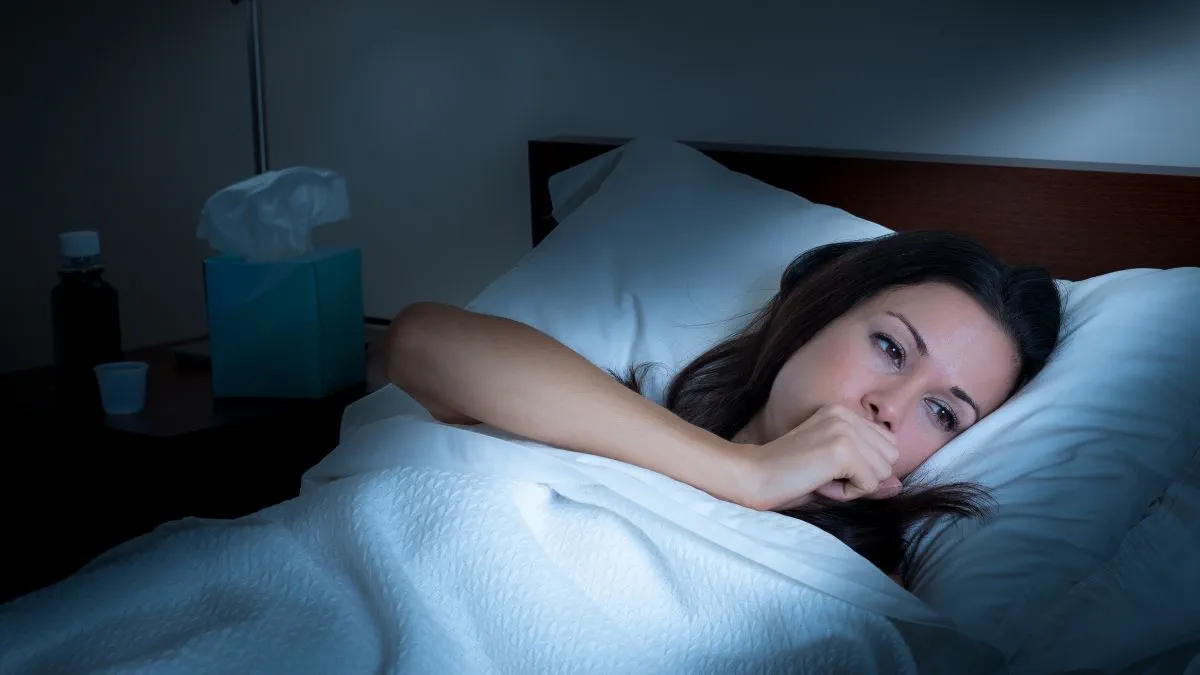 An adult sitting upright in bed at night with a hand on their chest, coughing, with a bedside lamp softly lighting the room