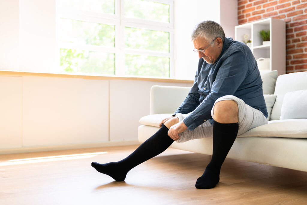 An older adult pulling on knee-high compression socks while sitting on the edge of a bed, with mild ankle swelling visible